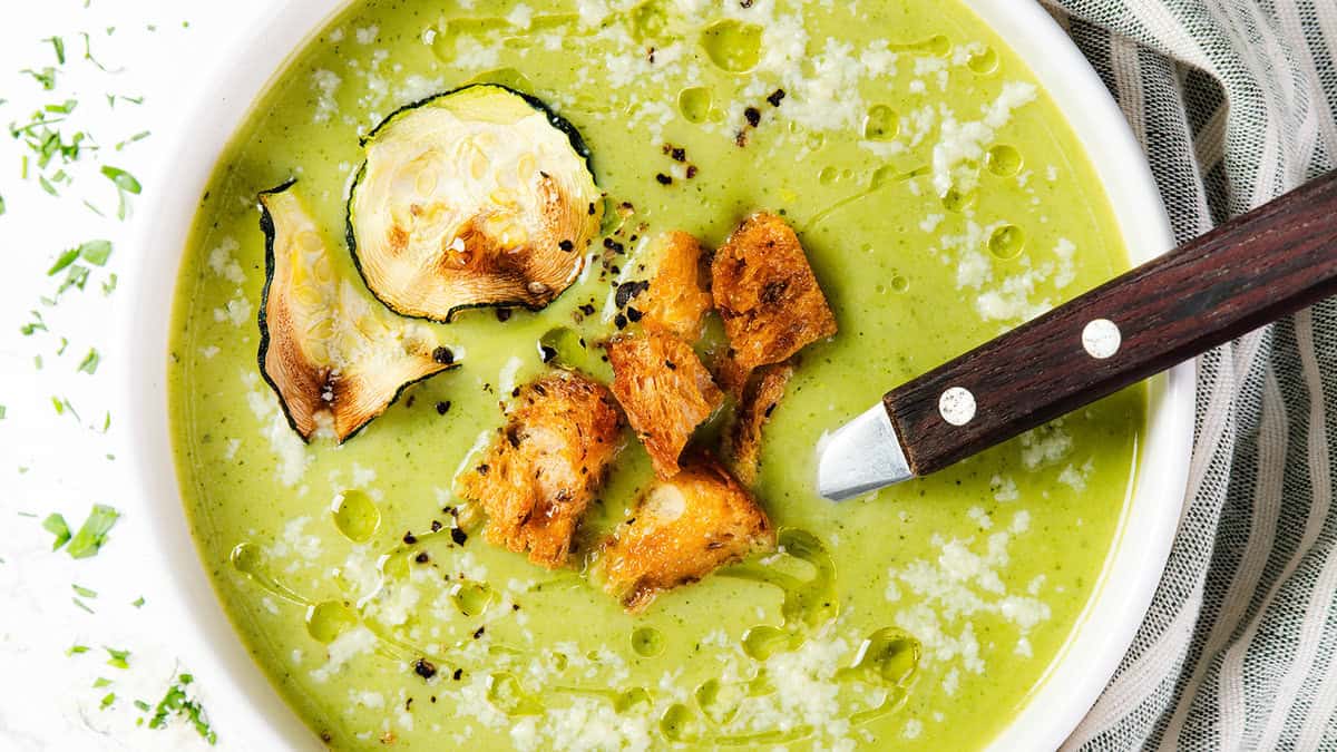 A green vegetable soup, perfect to fuel your day, is garnished with crispy zucchini slices and seasoned croutons in a white bowl. A wooden-handled spoon rests in the soup. Drizzled with olive oil and sprinkled with herbs, this healthy soup is accompanied by a striped napkin beside the bowl.