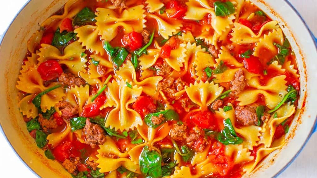 A large pot of hearty soup containing bow-tie pasta, ground beef, diced tomatoes, spinach, and a rich tomato-based broth. The ingredients are well mixed, creating a colorful and appetizing combination—perfect for anyone looking for delicious ground beef recipes.