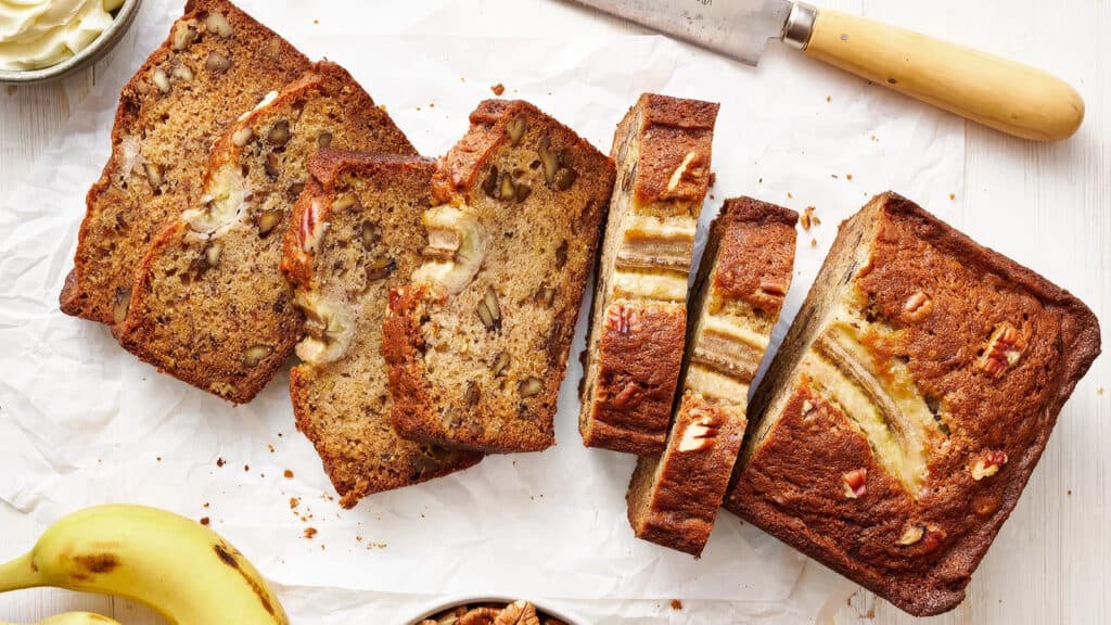 A sliced loaf of banana bread with nuts and banana pieces on top, laid out on parchment paper. A knife with a wooden handle rests nearby. A whole banana, a bowl of chopped nuts, and a dish with spread are also visible in the scene.