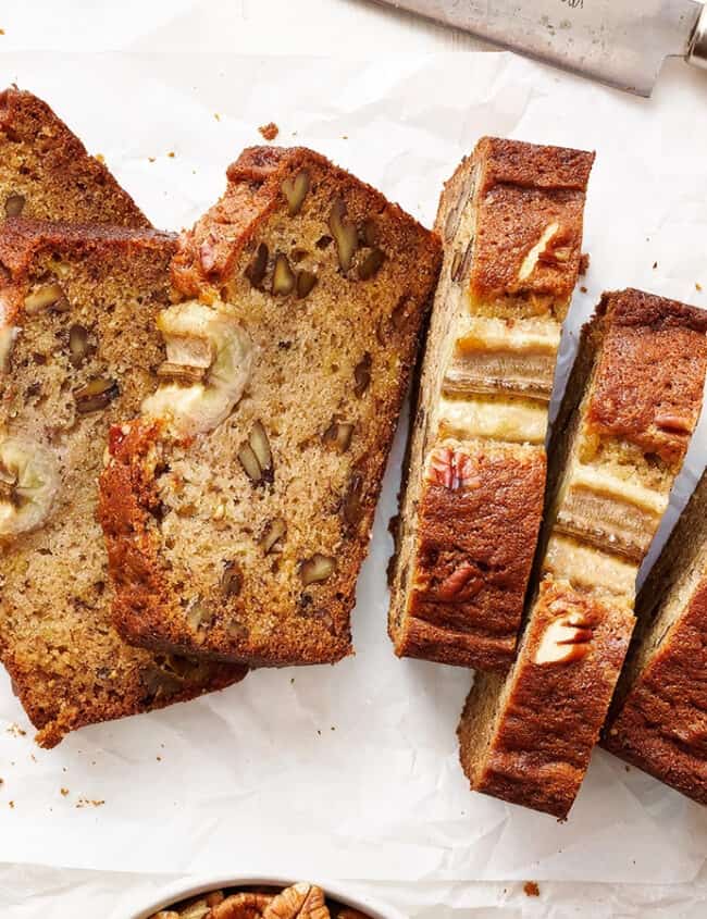 A sliced loaf of banana bread with nuts and banana pieces on top, laid out on parchment paper. A knife with a wooden handle rests nearby. A whole banana, a bowl of chopped nuts, and a dish with spread are also visible in the scene.