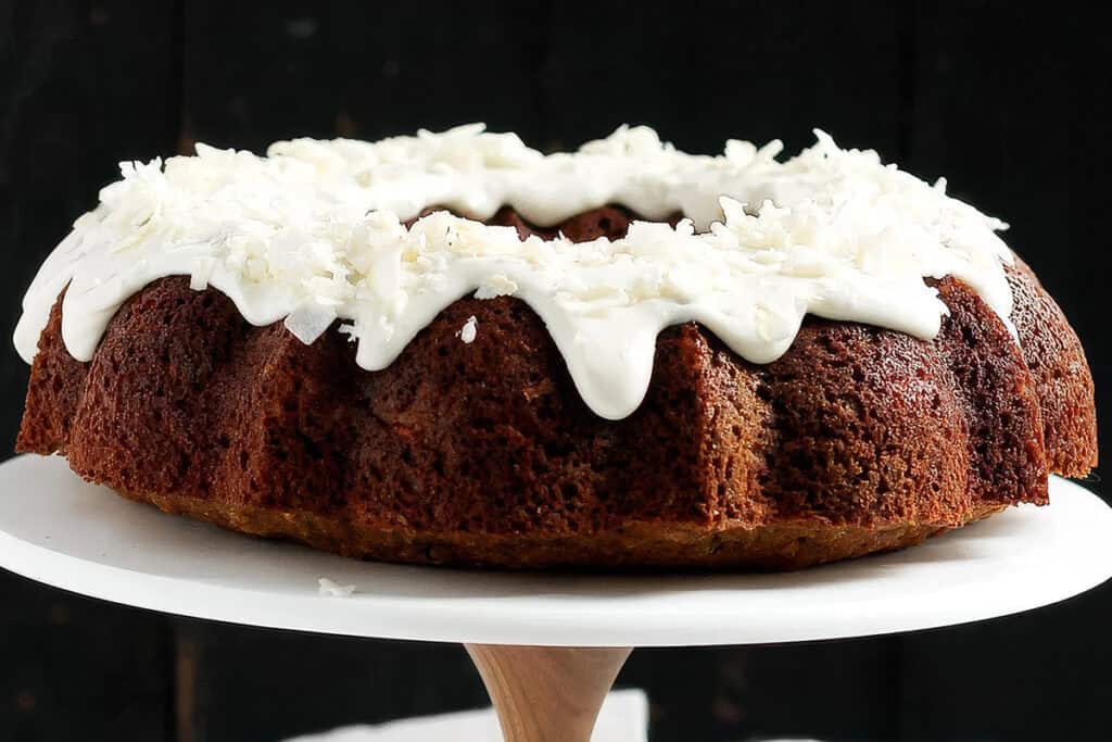 A Bundt cake topped with a thick layer of white frosting and garnished with shredded coconut, from the finest Bundt cake recipes. The cake is displayed on a white cake stand against a dark background.