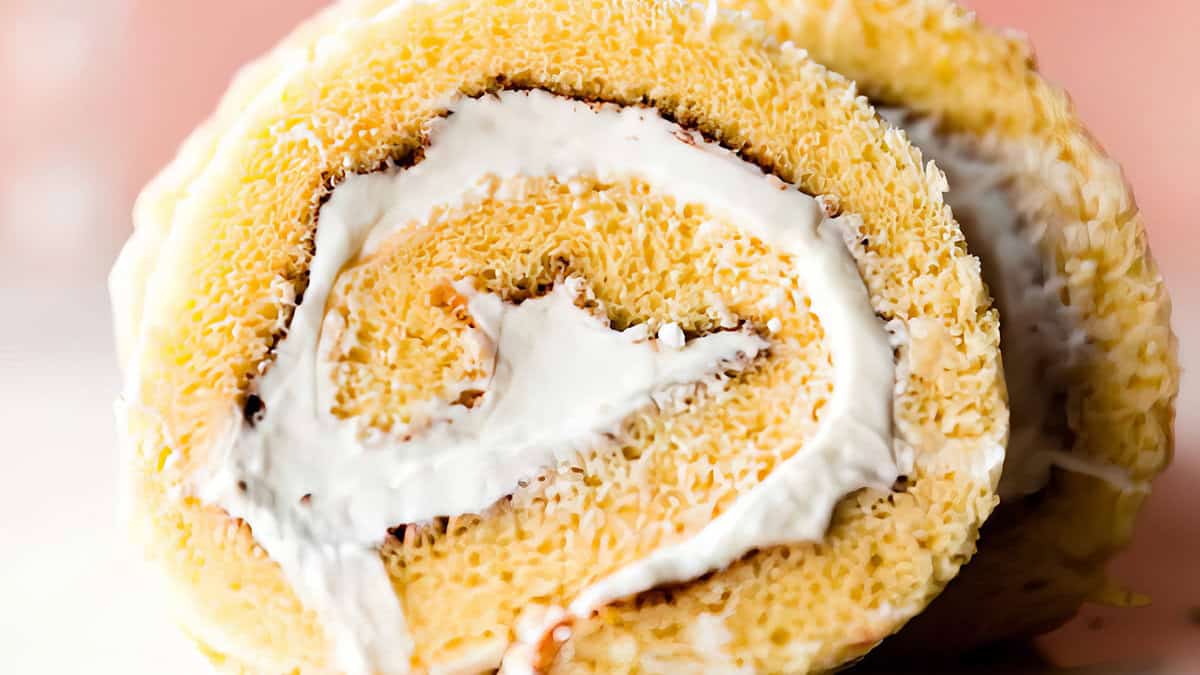 A close-up shot of a Swiss roll cake, featuring a golden-yellow sponge cake rolled with a white cream filling. The cake's spiral pattern is clearly visible, with the creamy filling contrasting against the sponge. The background is softly blurred.