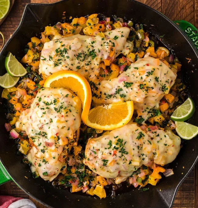 A skillet filled with baked chicken breasts topped with melted cheese, garnished with chopped parsley. This delightful chicken recipe is surrounded by colorful diced peppers and onions, with lime and orange slices for garnish. A red and white checkered cloth and various sides are visible.