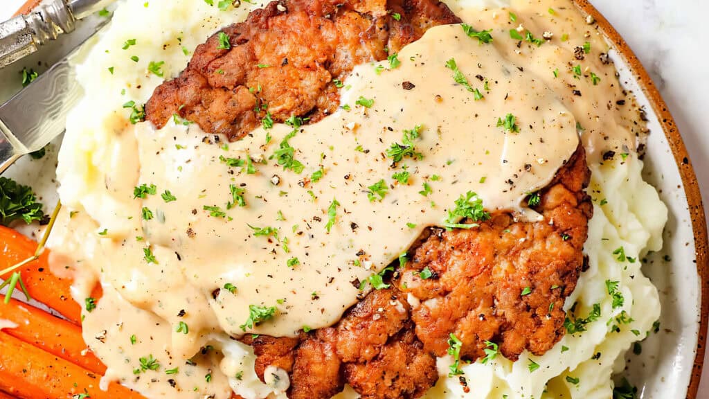 A plate of Southern fried chicken cutlets is served atop mashed potatoes and smothered in a creamy gravy. The dish is garnished with chopped parsley and freshly ground black pepper, with a side of glazed carrots visible in the corner.