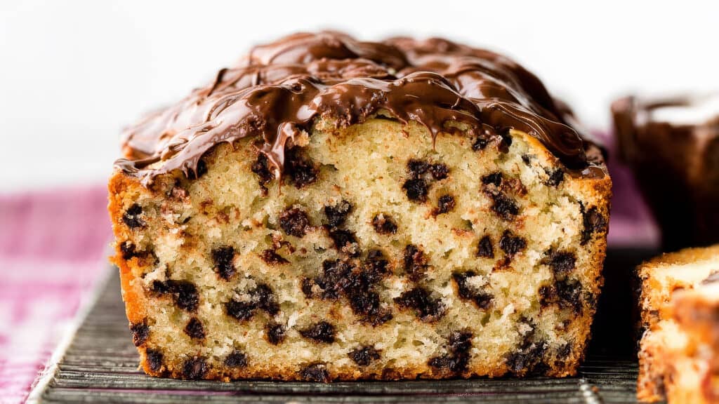 Close-up of a loaf of chocolate chip bread with a rich chocolate glaze on top. The loaf is sliced, revealing a moist interior dotted generously with chocolate chips, reminiscent of decadent loaf cakes. The background includes a blurred view of another loaf and a pink cloth.