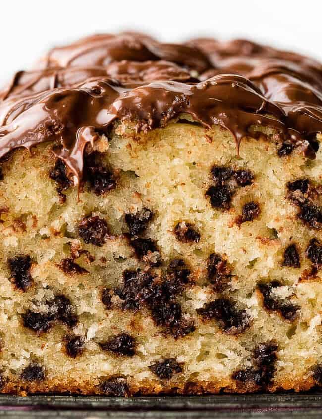 Close-up of a loaf of chocolate chip bread with a rich chocolate glaze on top. The loaf is sliced, revealing a moist interior dotted generously with chocolate chips, reminiscent of decadent loaf cakes. The background includes a blurred view of another loaf and a pink cloth.