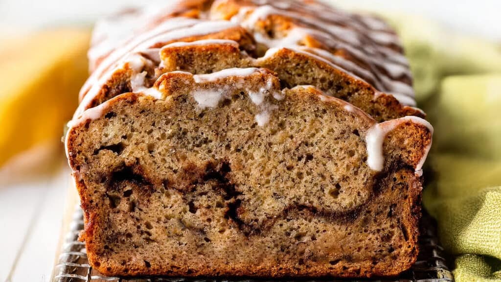 Close-up of a sliced loaf of banana bread with a swirl of cinnamon running through it, topped with a light drizzle of icing. The bread has a golden brown crust and a moist, dense texture. A yellow cloth is partially visible in the background, perfect for fans of homemade cakes.