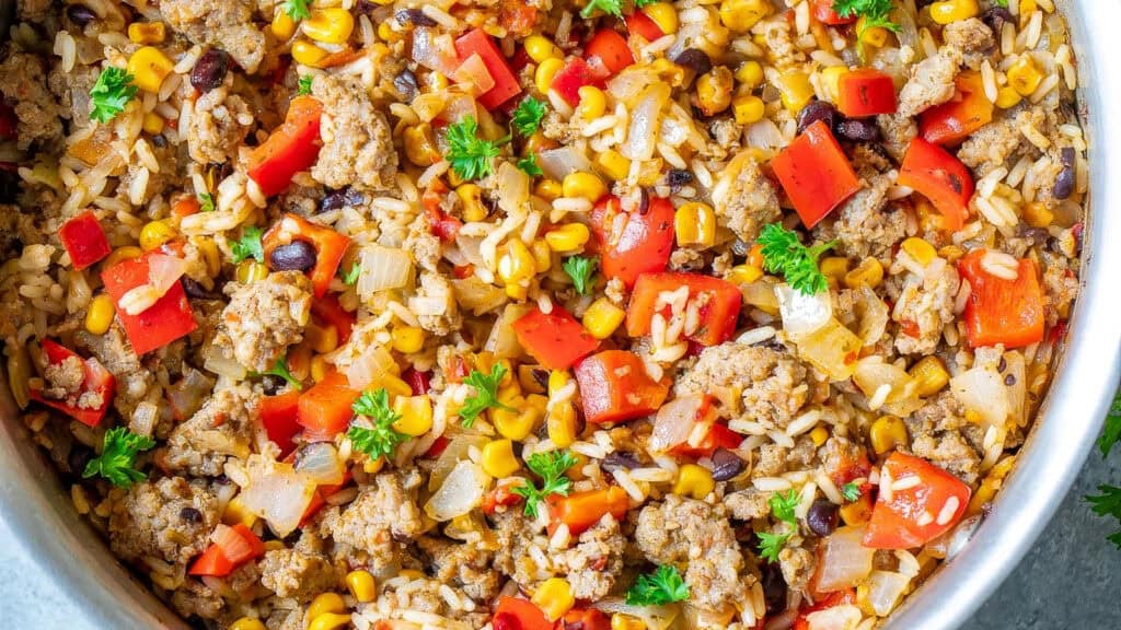 A vibrant and colorful dish from the list of popular ground beef recipes, featuring ground meat, rice, black beans, corn, diced red bell peppers, and chopped onions seasoned with herbs and spices, garnished with fresh parsley. The ingredients are mixed and cooked together in a large skillet.