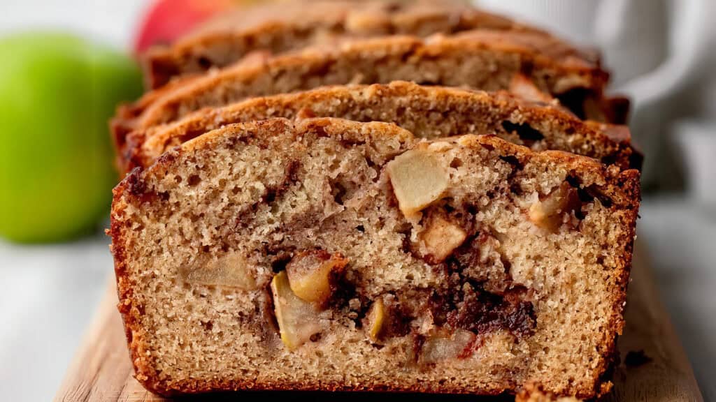 Close-up image of sliced apple cinnamon bread. The moist homemade loaf cake features visible chunks of apple and swirls of cinnamon throughout. It sits on a wooden cutting board with a green apple blurred in the background. The texture looks soft and inviting.