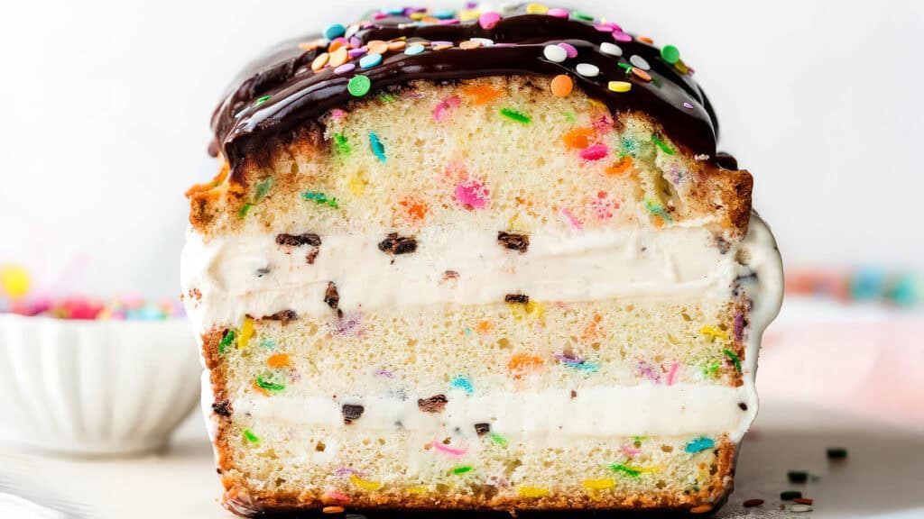 Close-up of a slice of confetti cake with a rich layer of chocolate frosting on top and colorful sprinkles. This delightful dessert features two layers of white frosting sandwiched between moist, speckled cake layers, giving a vibrant and festive appearance.