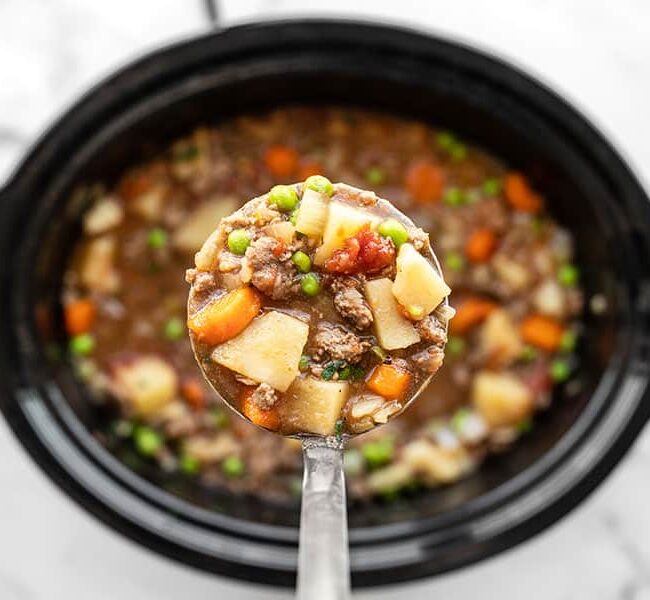 A ladle filled with hearty beef stew containing chunks of meat, potatoes, carrots, and peas. The ladle is held over a crockpot filled with more of the same stew, showcasing a delicious mix of ingredients in a rich broth.