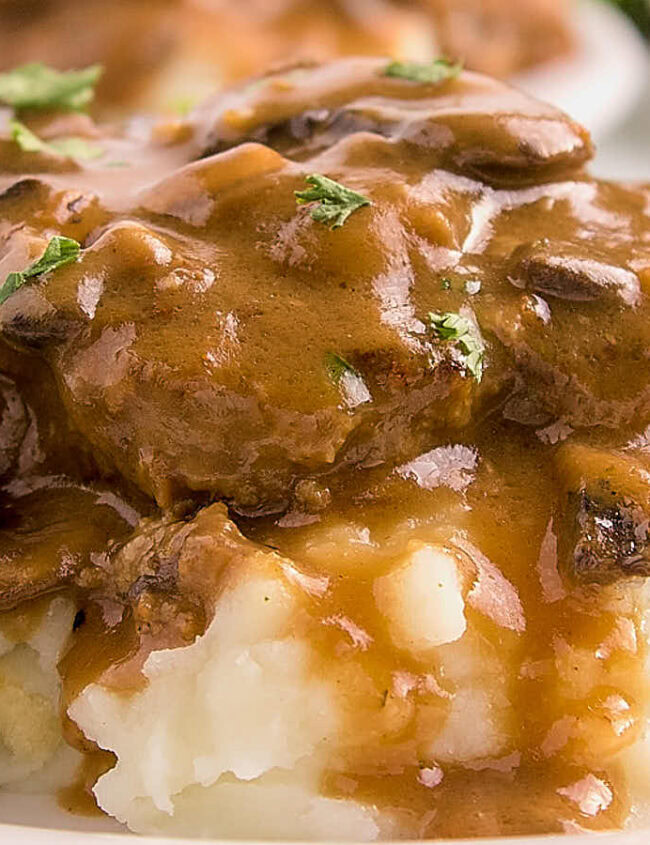 A plate of mashed potatoes is topped with a hearty brown gravy, filled with pieces of sautéed mushrooms and garnished with chopped parsley. In the background, there is a serving of asparagus.