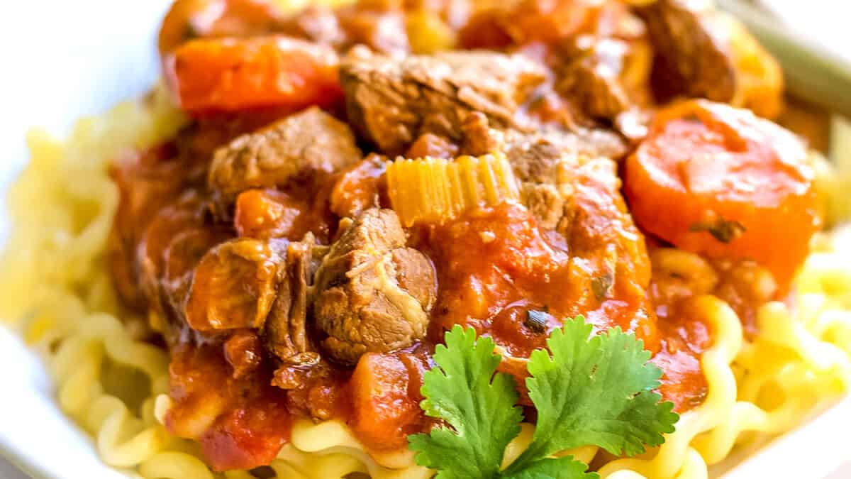 A close-up of a plate of pasta topped with a hearty meat and tomato sauce, epitomizing comfort food. Visible ingredients include chunks of meat, sliced carrots, and celery pieces, garnished with a sprig of fresh cilantro. The curly yellowish pasta underneath completes this delectable recipe.