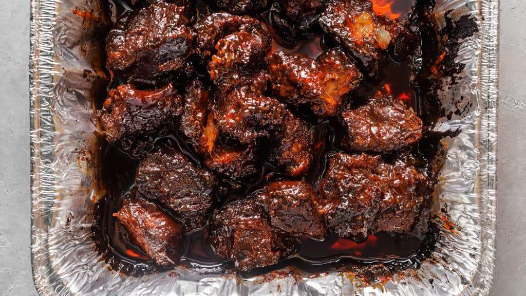 An overhead view of a foil tray filled with glazed and caramelized burnt ends. The meat pieces are dark and glossy, indicating a rich and flavorful sauce coating—a true testament to Southern recipes. The tray sits on a light-colored surface.