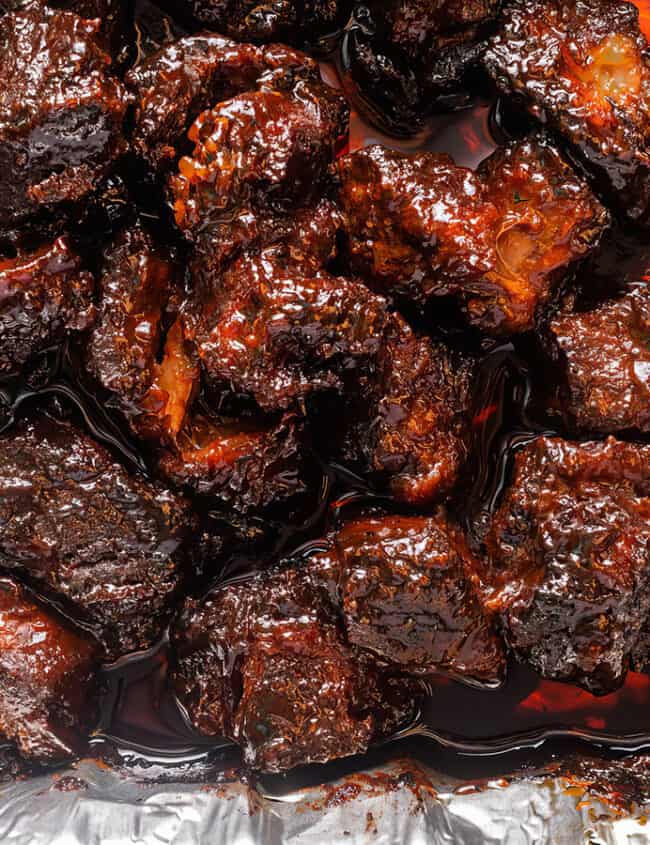 An overhead view of a foil tray filled with glazed and caramelized burnt ends. The meat pieces are dark and glossy, indicating a rich and flavorful sauce coating—a true testament to Southern recipes. The tray sits on a light-colored surface.