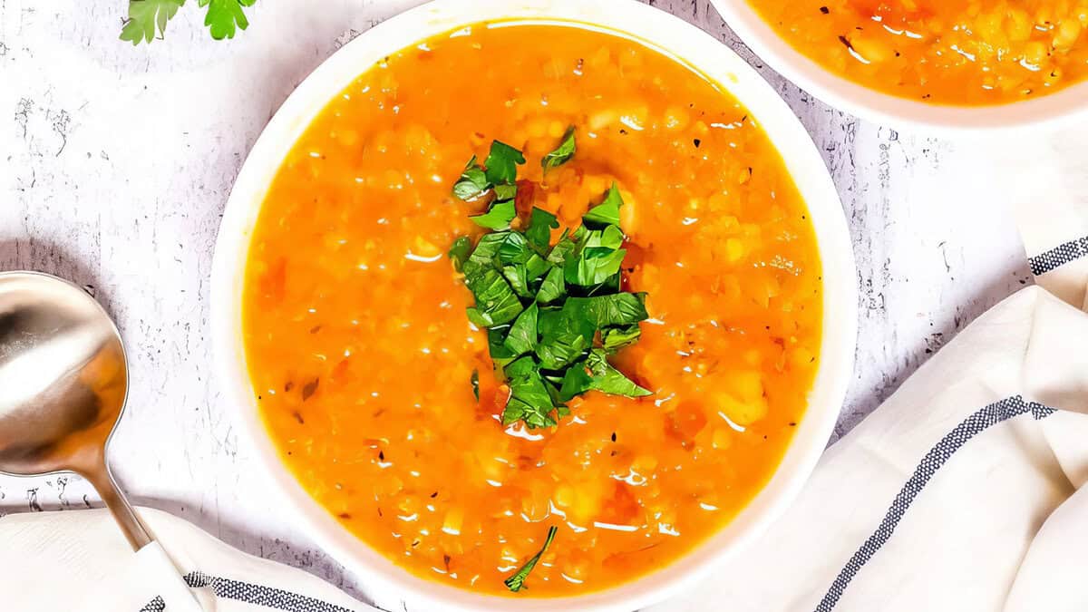 A bowl of vibrant orange soup, renowned for its healthy ingredients, is garnished with fresh green herbs and set on a white table. A silver spoon rests to the side, and a white cloth with dark stripes partially covers the table. The background is light and slightly textured.