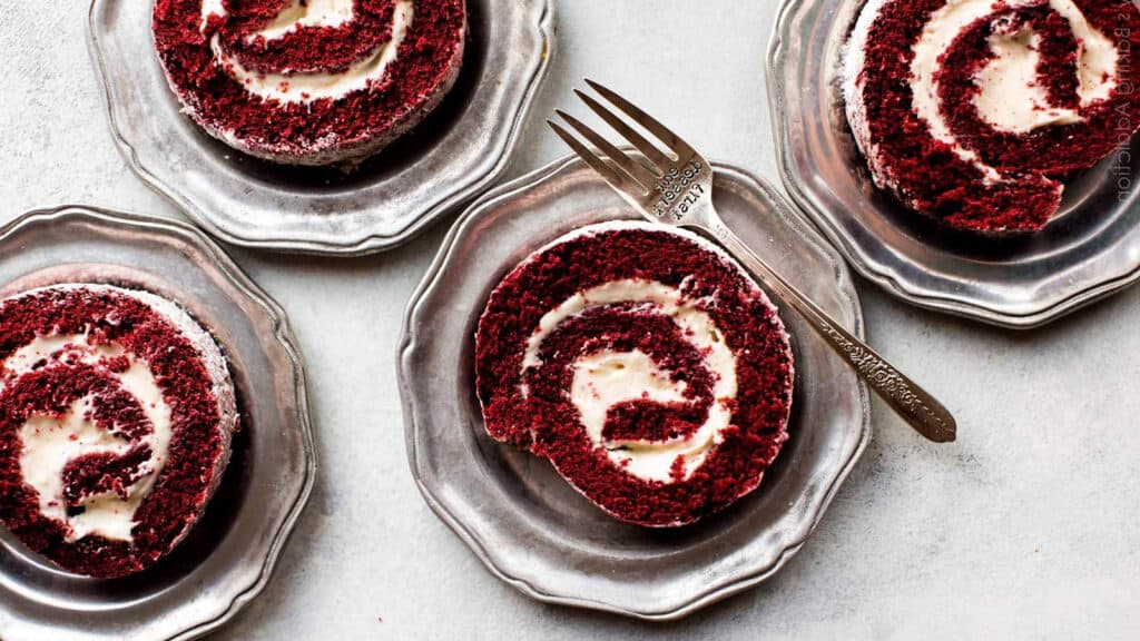 Three neatly sliced red velvet cake rolls with cream cheese filling are placed on silver plates, accompanied by a fork. The vibrant red color of the cake contrasts with the creamy white filling, creating an elegant and appetizing presentation.