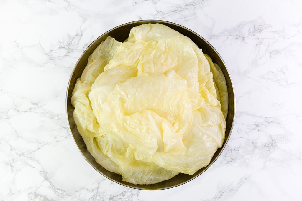 A bowl filled with large, pale yellow, translucent napa cabbage leaves reminiscent of those used in Romanian Cabbage Rolls sits on a white marble countertop. The leaves appear slightly wilted or steamed.