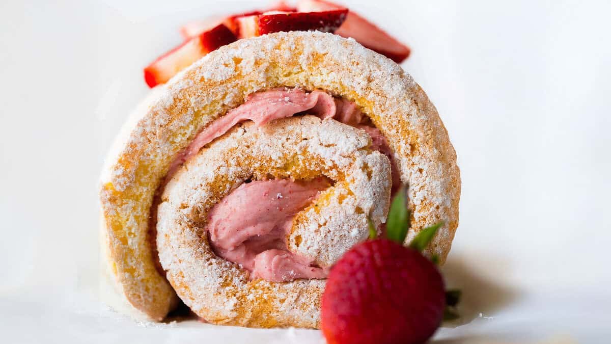 Close-up of a sliced Swiss roll cake filled with pink strawberry cream, topped with fresh strawberry slices, and dusted with powdered sugar. A whole red strawberry with green leaves is placed in front of the cake. The background is white and blurred.