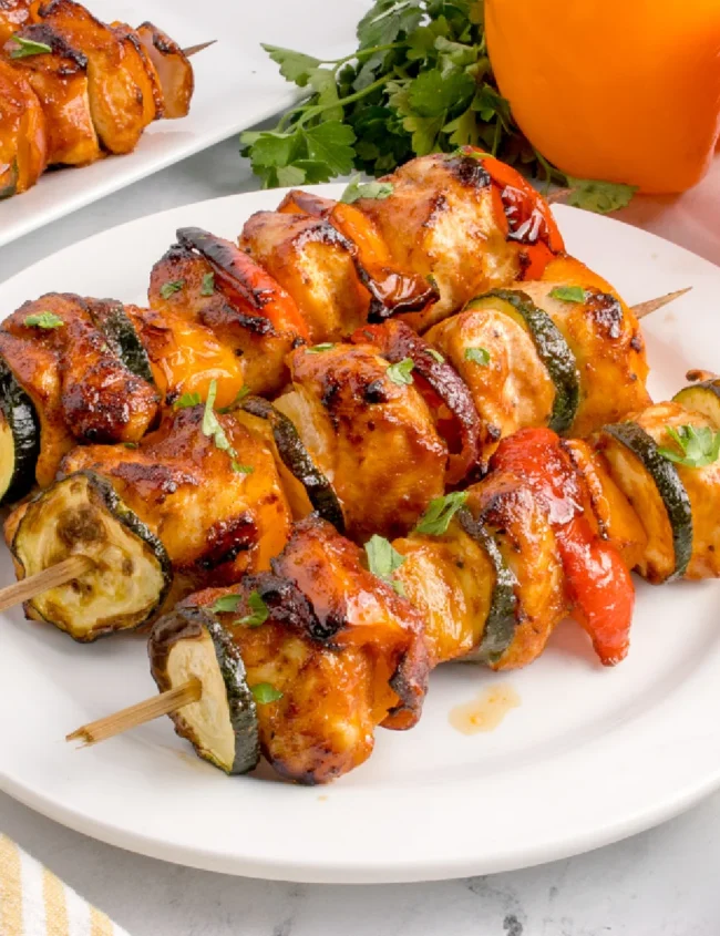 Close-up of grilled chicken and vegetable skewers on a white plate, perfect for air fryer recipes. The skewers include chicken, zucchini, and red and yellow bell peppers, garnished with fresh herbs. In the background, whole peppers and parsley are visible.