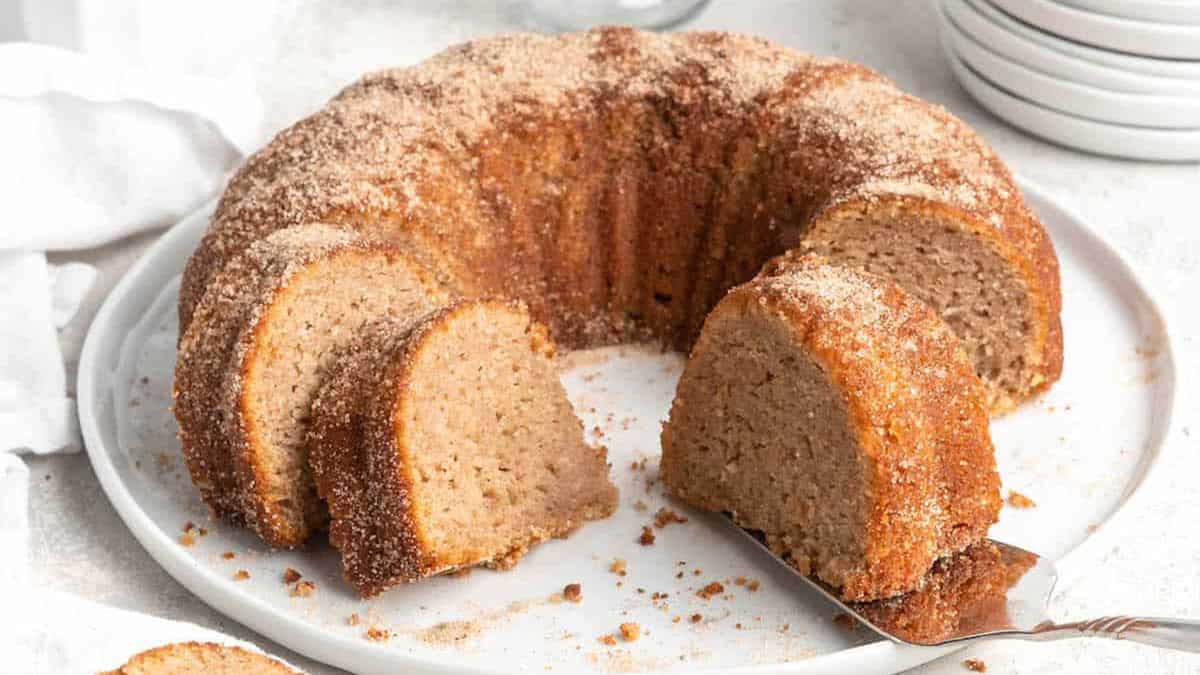 A cinnamon sugar bundt cake with a few slices cut and arranged on a white plate, showcasing the warm tones of the mix. The cake has a light brown color and a sugar-dusted surface, perfect for those seeking delightful Bundt Cake recipes. Plates and a napkin complete the cozy scene.