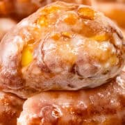 A tantalizing close-up of apple fritters highlights their shiny, sugary glaze and textured surface, with visible pieces of apple and cinnamon. These apple fritters are piled together, offering a warm and inviting bakery treat that's impossible to resist.