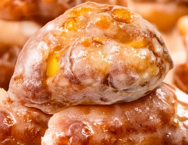 A tantalizing close-up of apple fritters highlights their shiny, sugary glaze and textured surface, with visible pieces of apple and cinnamon. These apple fritters are piled together, offering a warm and inviting bakery treat that's impossible to resist.