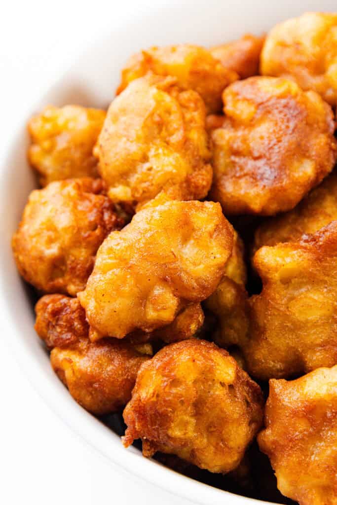 A close-up of golden brown apple fritters piled in a white bowl. The fritters have a crispy, textured surface, hinting they might be freshly fried from a cherished fritter recipe.