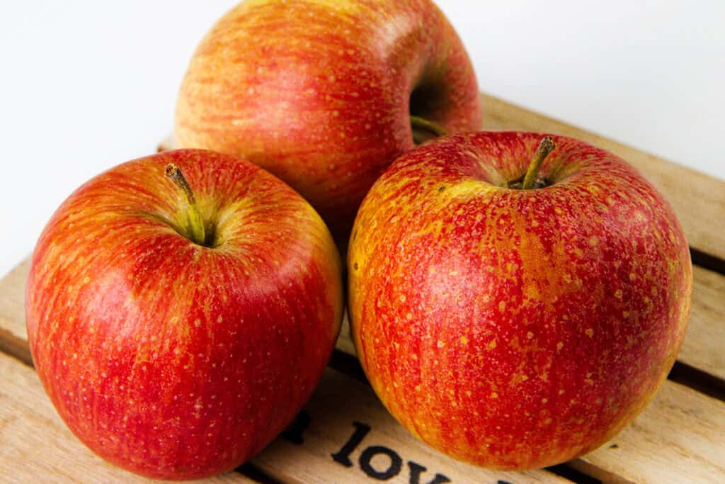 Three red and yellow apples with speckled skin sit on a wooden surface, their shiny appearance suggesting they are fresh and ripe, perfect for creating a delightful apple dessert.