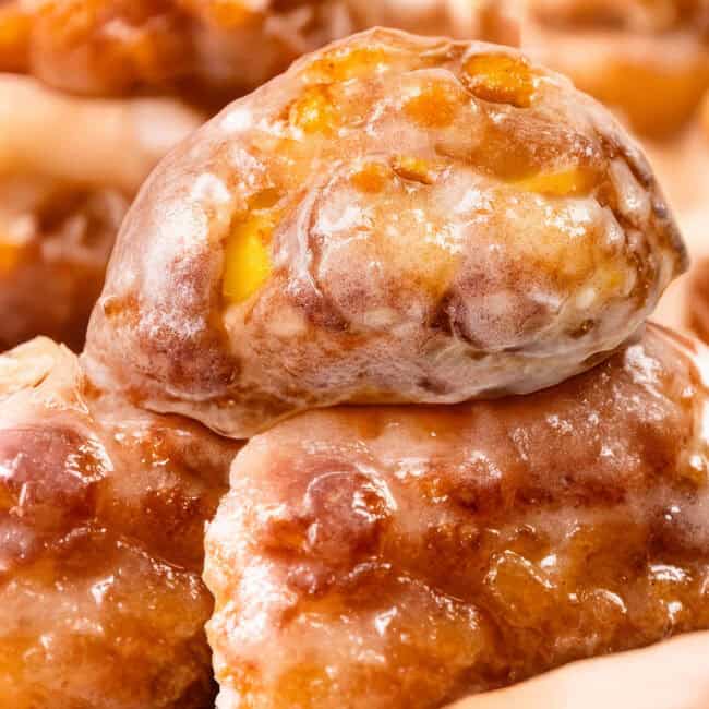 Close-up of delectable apple fritters stacked together. These golden brown delights boast a shiny glaze and visible chunks of apple, offering a textured and appetizing appearance.