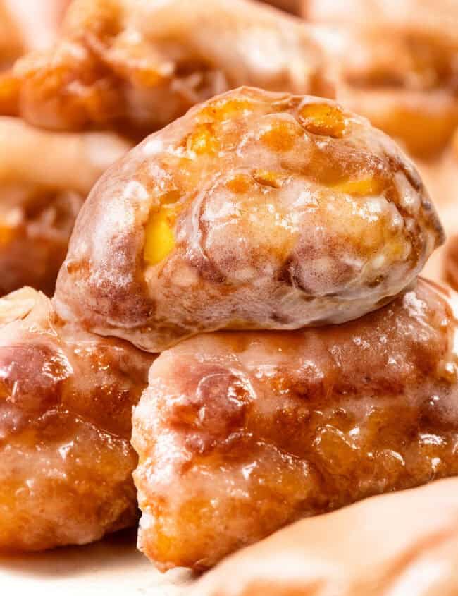 Close-up of delectable apple fritters stacked together. These golden brown delights boast a shiny glaze and visible chunks of apple, offering a textured and appetizing appearance.