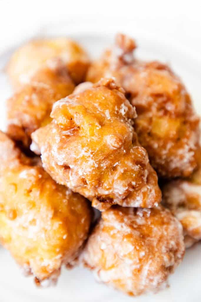 Close-up of a pile of glazed apple fritters, the perfect apple dessert. These sweet pastries are golden brown, crispy, and slightly irregular in shape, with a shiny glaze coating their surface. They rest invitingly on a white plate.