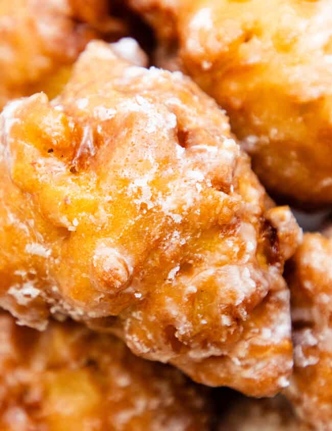 Close-up of several golden-brown, glazed apple fritters stacked on a plate. With their crispy, irregular texture and visible pieces of apple, these delightful treats are a must-try in dessert recipes, lightly drizzled with white glaze for the perfect finish.