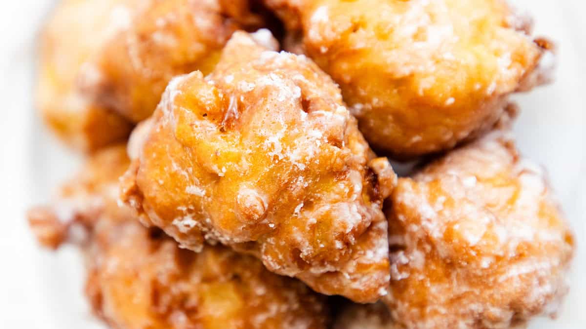 Close-up of several golden-brown, glazed apple fritters stacked on a plate. With their crispy, irregular texture and visible pieces of apple, these delightful treats are a must-try in dessert recipes, lightly drizzled with white glaze for the perfect finish.