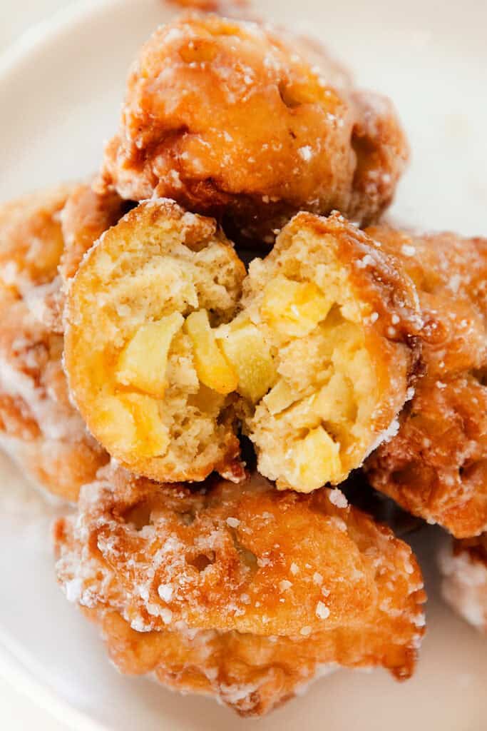 A close-up of several glazed apple fritters stacked on a white plate. The top fritter is broken open, revealing pieces of apple inside the dough. These delectable apple fritters boast a shiny, sweet glaze that enhances the golden-brown delight.