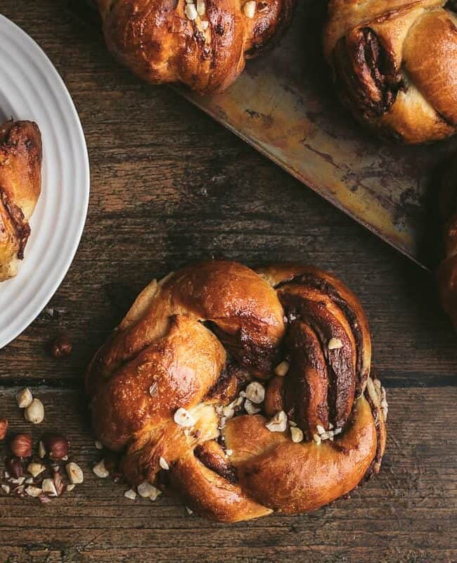 A rustic wooden table hosts a delightful scene of cinnamon rolls topped with crushed nuts, elegantly arranged on a metal tray and a white plate—a sweet escape from sticky situations.