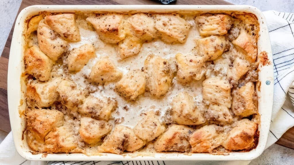 A Southern delight, this baked casserole features golden-brown biscuits atop a creamy sausage gravy. Perfectly set on a wooden board and partially covered by a white and black striped cloth, it captures the heart of comforting Southern cuisine.