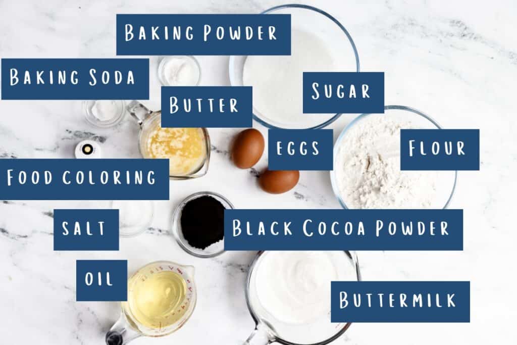 Image of baking ingredients on a white surface, labeled for creating a Black Velvet Cake: Baking Powder, Baking Soda, Butter, Sugar, Eggs, Flour, Food Coloring, Salt, Black Cocoa Powder, Oil, Buttermilk. Each ingredient is neatly arranged in its own bowl or container.