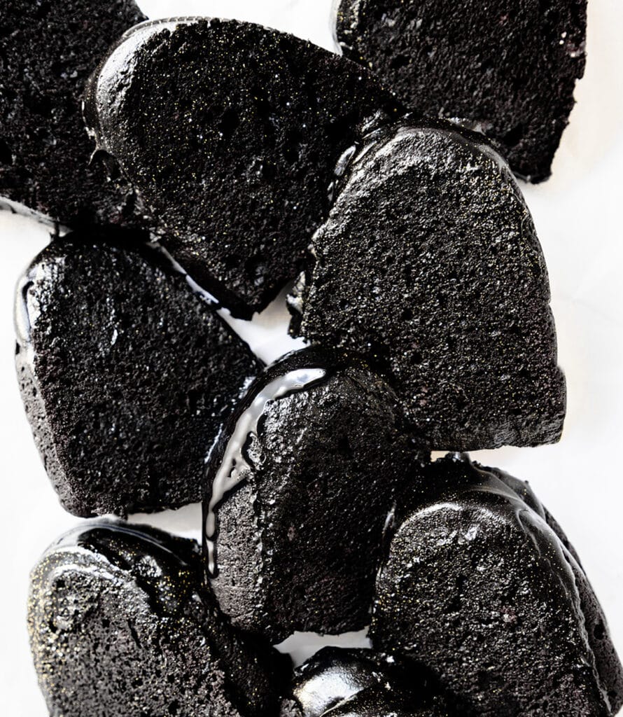 Close-up of glossy black slices, reminiscent of Black Velvet Cake, coated with a shiny glaze. The texture appears dense and moist, with pieces arranged haphazardly on a white surface.