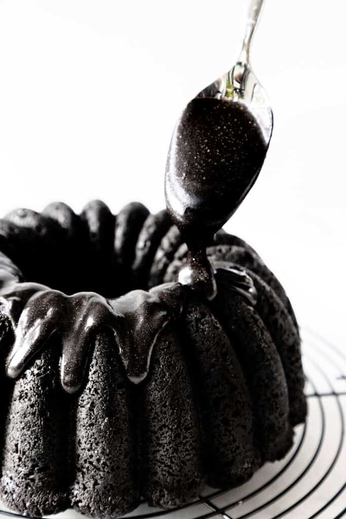 A dark, glossy Black Velvet Cake sits on a cooling rack. A spoon drizzles thick, black glaze over the cake, enhancing its rich texture and adding a shiny finish. The background is white, highlighting the dessert's luxurious appearance.