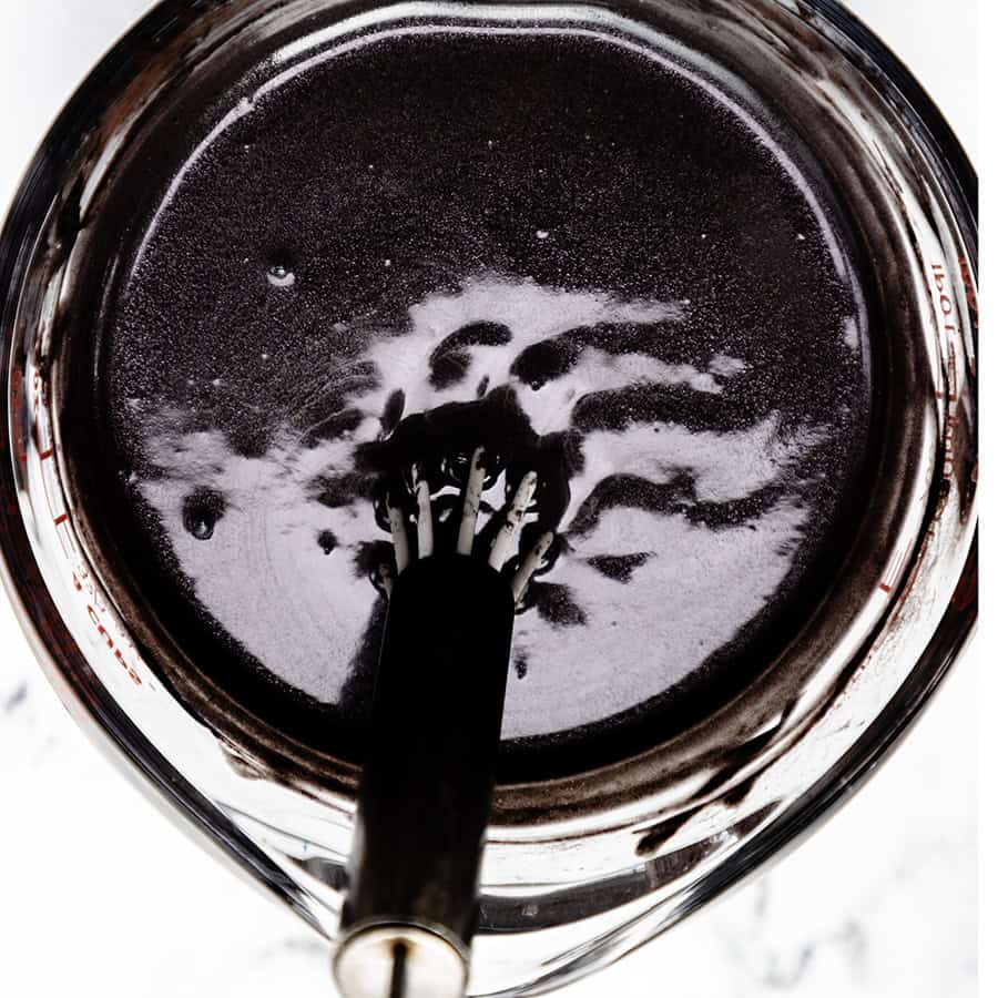 A close-up of a whisk in a bowl containing a dark, glossy liquid, reminiscent of Black Velvet Cake batter. The mixture appears rich and smooth, with reflections on the surface. The bowl is set on a light-colored surface, highlighting the luxurious allure of the dessert in progress.