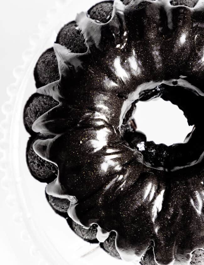 Top view of a glossy black velvet bundt cake with a shiny, metallic sheen on a white background. The texture appears smooth and sleek, highlighting the cake’s intricate curved design.