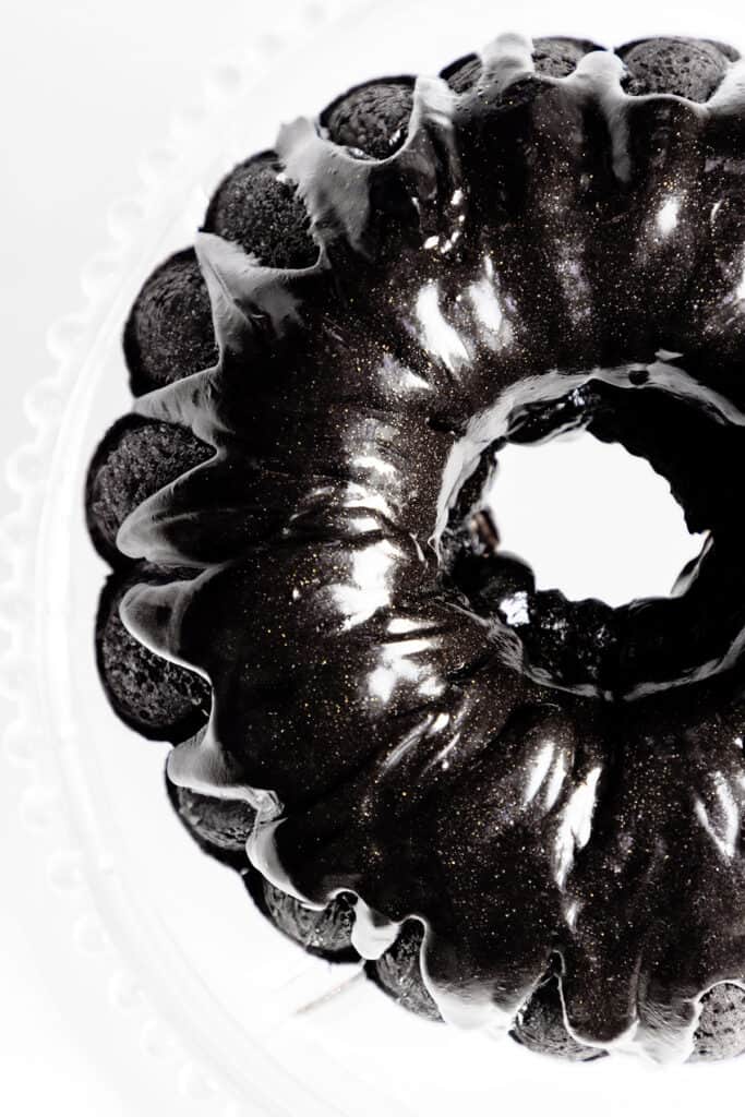 Top view of a glossy black velvet bundt cake with a shiny, metallic sheen on a white background. The texture appears smooth and sleek, highlighting the cake’s intricate curved design.