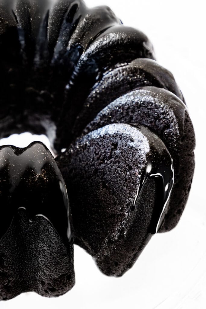 Close-up of a glossy, Black Velvet bundt cake with a shiny glaze. The cake's rich texture and curved ridges are highlighted in the lighting, enhancing its decadent appearance.