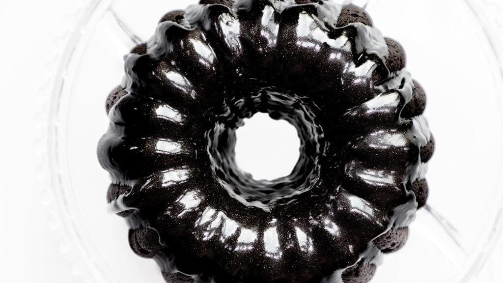 A show-stopping black bundt cake with a shiny glaze sits on a clear glass plate, viewed from above. The dark, glossy surface contrasts with the bright background, highlighting its wavy, circular shape. This stunning dessert is simple to make at home and perfect for any cake lover.