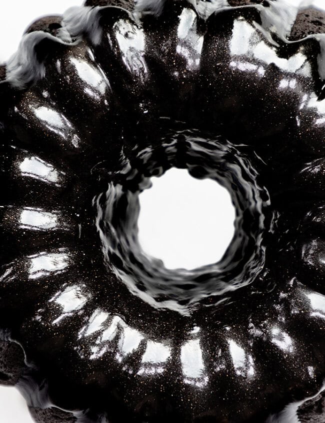 A show-stopping black bundt cake with a shiny glaze sits on a clear glass plate, viewed from above. The dark, glossy surface contrasts with the bright background, highlighting its wavy, circular shape. This stunning dessert is simple to make at home and perfect for any cake lover.