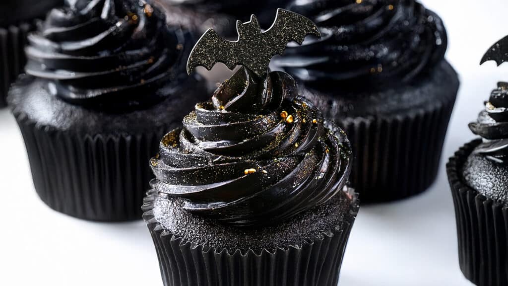 Black cupcakes with dark, glossy frosting are topped with bat-shaped decorations. Gold specks are scattered on the frosting, giving these Halloween treats a spooky, festive appearance that's perfect for the season.