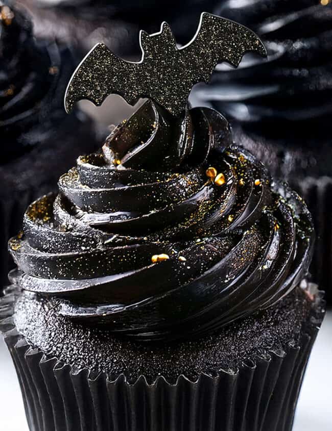 Black cupcakes with dark, glossy frosting are topped with bat-shaped decorations. Gold specks are scattered on the frosting, giving these Halloween treats a spooky, festive appearance that's perfect for the season.