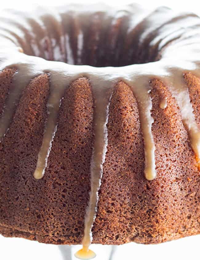 A luscious bundt cake with smooth, glossy icing drizzled on top sits elegantly on a glass platform. The golden-brown cake is perfectly complemented by the caramel-like icing, which delicately drips down its sides, creating a mouthwatering spectacle.