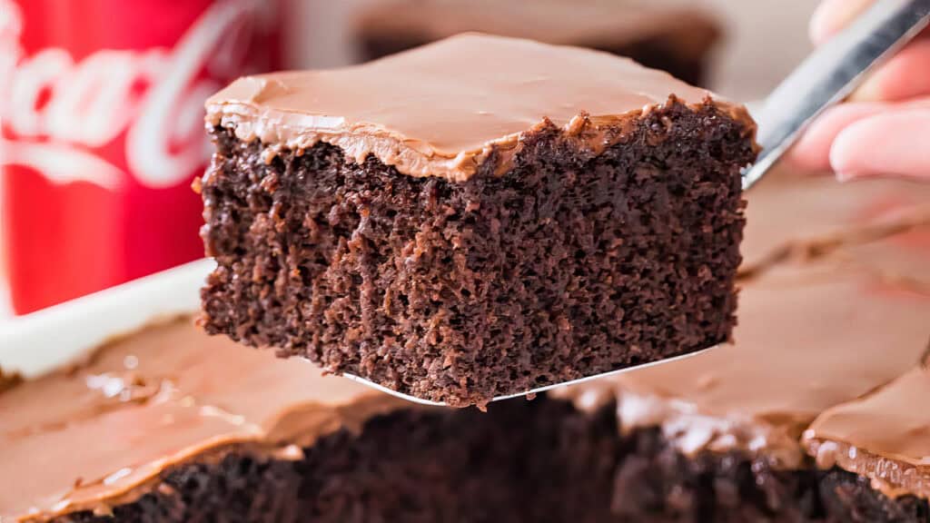 A close-up of a slice of moist, chocolate cake with a smooth chocolate frosting, being lifted from a tray. A red soda can is partially visible in the background.